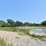 River flowing on the right. Sandy beach on the left. Trees in the background.