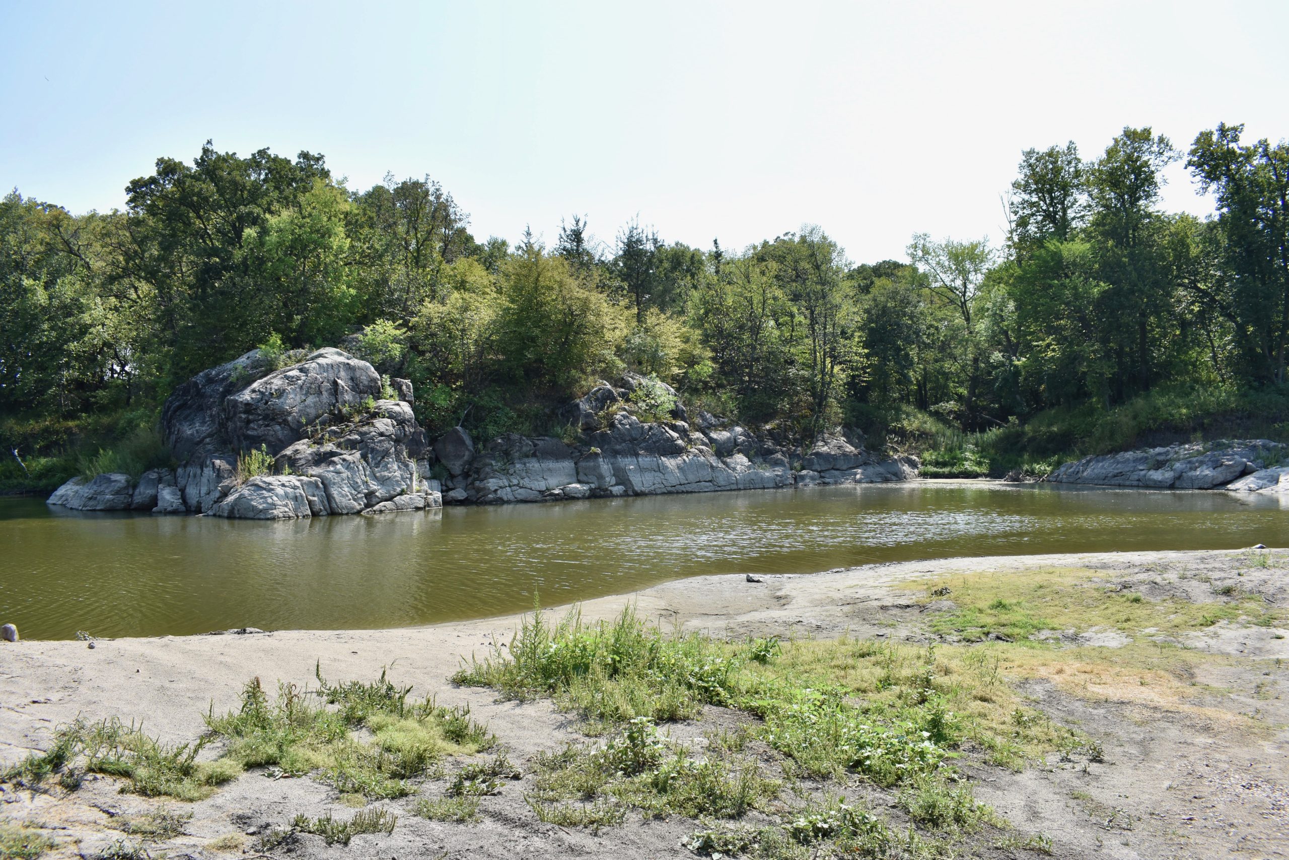 Rocky cliffs and trees in background. River and sandy beach in foreground.